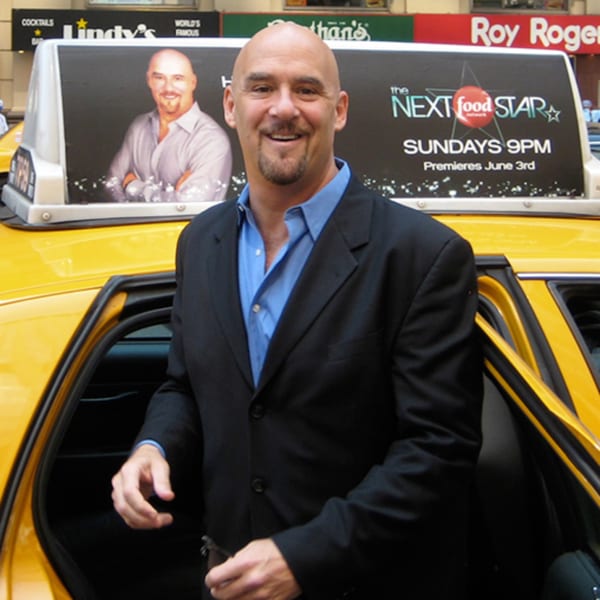 A smiling bald man in a suit stands next to a yellow taxi with a "Next Food Star" ad.