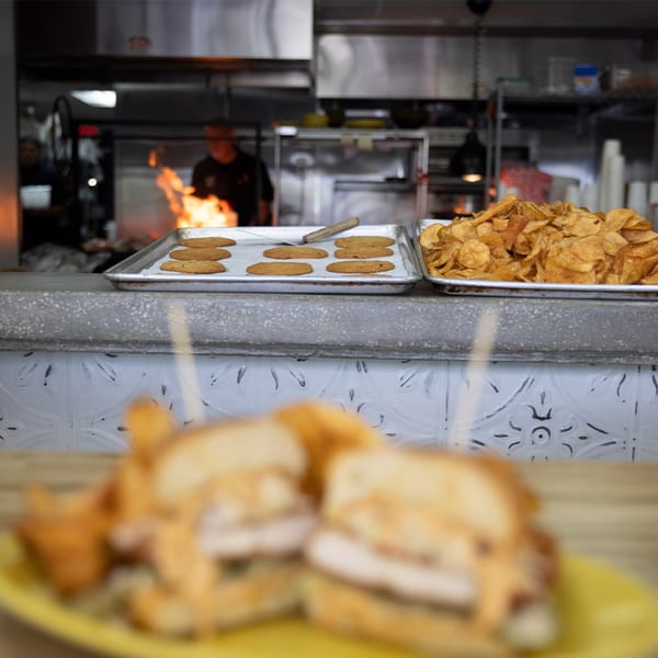 A chef cooks over a flaming grill, with cookies and chips on the counter, and blurred sandwiches in the foreground.