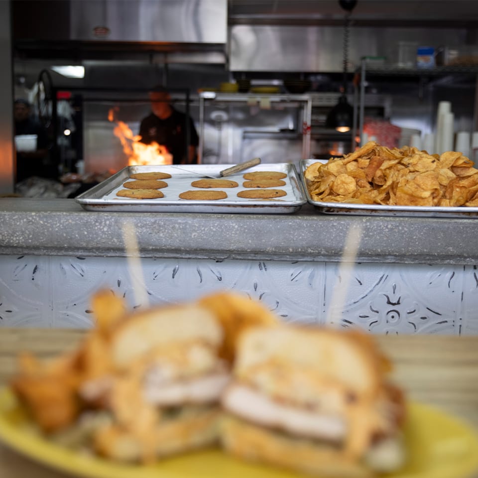 A chef cooks over a flaming grill, with cookies and chips on the counter, and blurred sandwiches in the foreground.