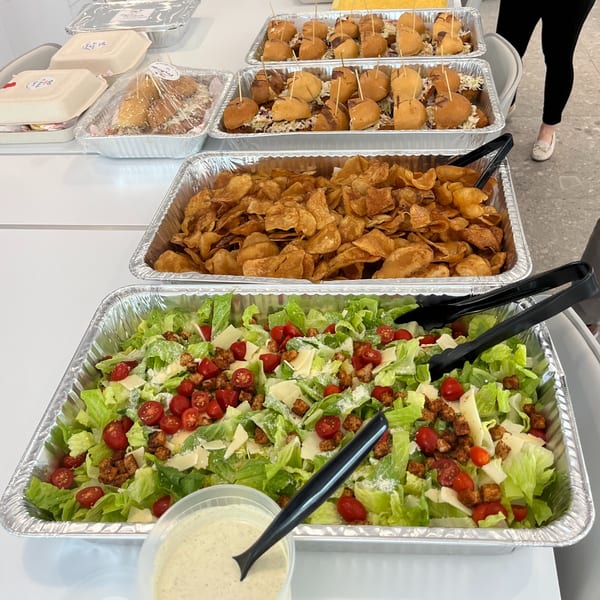 Catering spread with chicken sliders, chips, and Caesar salad in foil trays, with a person's legs in the background.