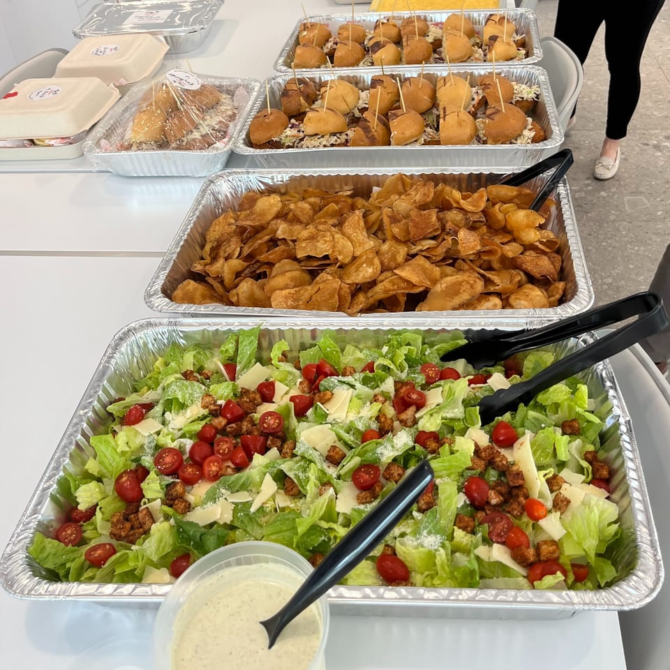 Catering spread with chicken sliders, chips, and Caesar salad in foil trays, with a person's legs in the background.